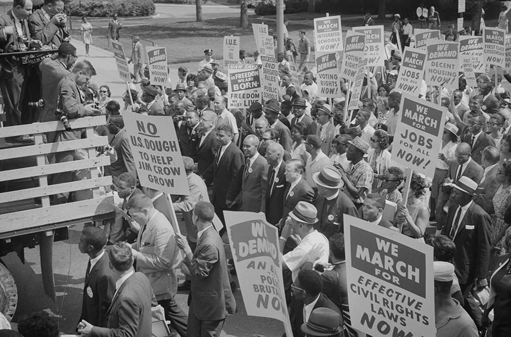 Civil rights leaders, including Martin Luther King, Jr., are surrounded by crowds carrying signs during the 1963 March on Washington.